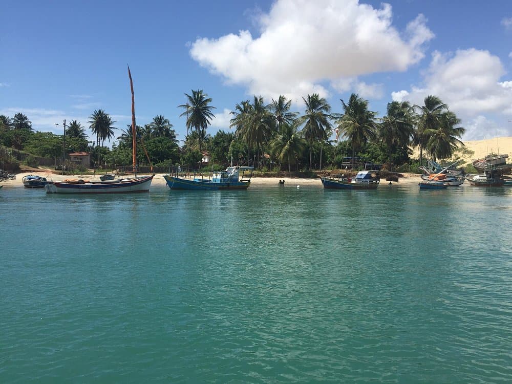 Barcos de pesca ancorados na água turquesa do estuário de Mundaú, com coqueiros e duna ao fundo